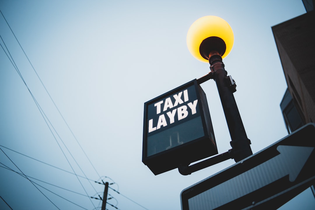 En tänd "Taxi Layby"-skylt hänger från en stolpe toppad med ett glödande sfäriskt ljus mot en mulen himmel. Kraftledningar sträcker sig över scenen, förstärker den urbana stämningen, samtidigt som den antyder en taxitjänst som erbjuder snabba och säkra resor med professionella chaufförer i närheten.